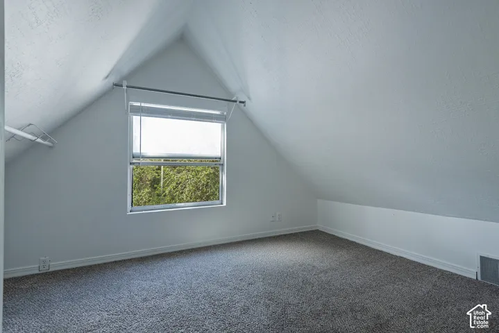 Attic space featuring sloped ceilings, a window with blinds, and gray carpeting