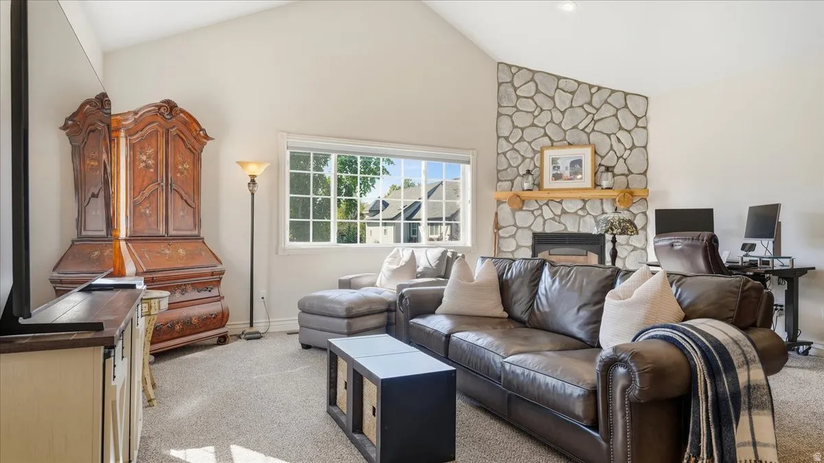 Vaulted ceiling, stone-clad fireplace with wood mantel, large window with grid pattern, and neutral carpeting