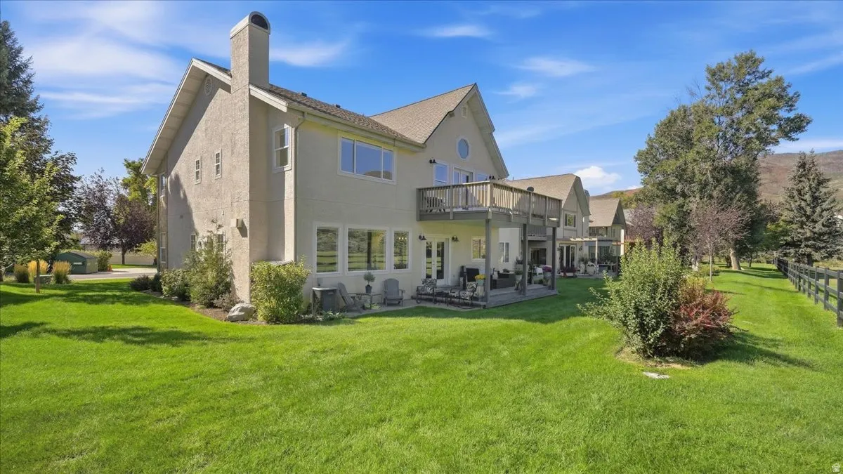 Expansive rear yard with a manicured lawn, multiple mature trees, a two-story stucco exterior, upper-level deck, and lower-level patio