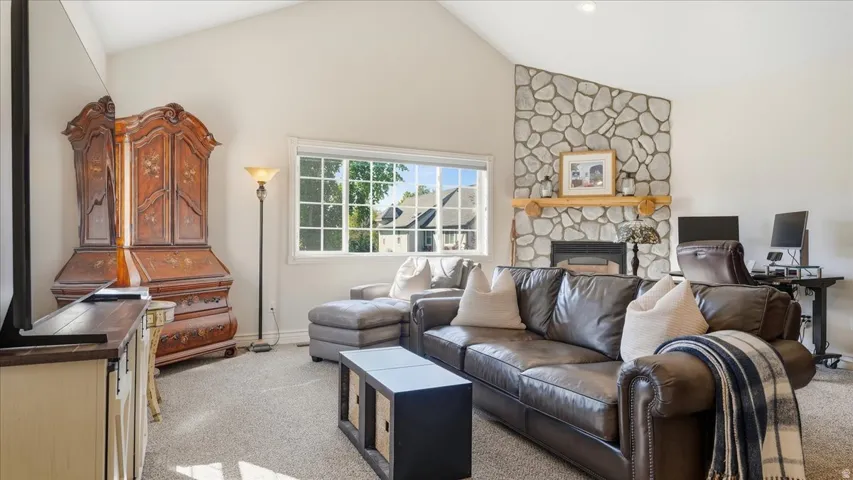 Vaulted ceiling, stone-clad fireplace with wood mantel, large window with grid pattern, and neutral carpeting
