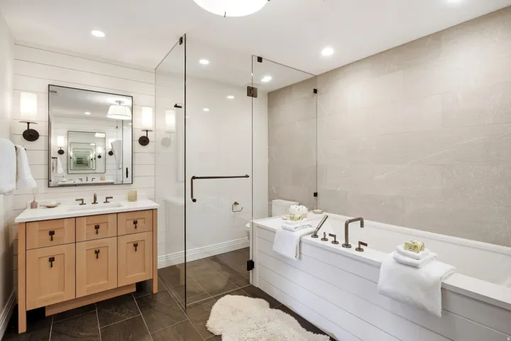Bathroom featuring a freestanding soaking tub, glass-enclosed shower, wood vanity with a white countertop, wall-mounted sconces, and dark tile flooring