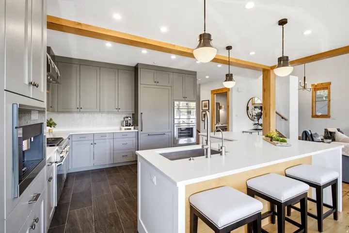 Kitchen featuring light gray cabinetry, built-in appliances, a center island with an undermount sink, pendant lighting, and exposed wood beams
