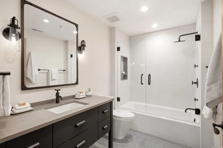 Bathroom featuring a vanity with an integrated sink, a large framed mirror, industrial-style wall sconces, a shower-over-tub with a glass enclosure, and matte black fixtures
