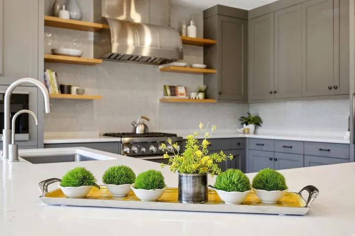 White countertop with integrated sink, stainless steel faucet, grey cabinetry, stainless steel range hood, and open wood shelving