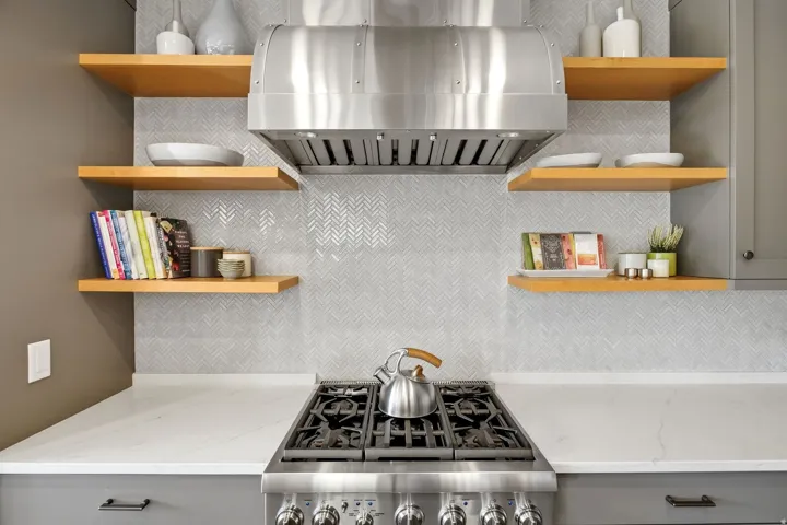 Stainless steel range hood, herringbone tile backsplash, wood-finish open shelving, white countertop, and gray lower cabinetry
