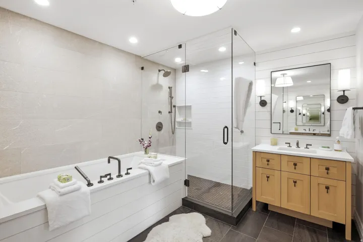 Glass-enclosed shower, built-in bathtub with dark fixtures, light wood-finish vanity, rectangular undermount sink, and wall-mounted sconces