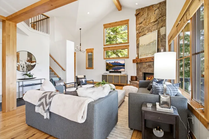 Vaulted ceiling with exposed wood beams, stone fireplace with wood mantel, wood-finish flooring, large windows with wood trim, and a staircase with a metal railing