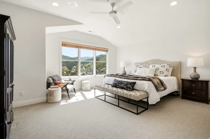 Vaulted ceiling with recessed lighting, ceiling fan, expansive window with natural wood blinds, upholstered headboard, and plush carpeting