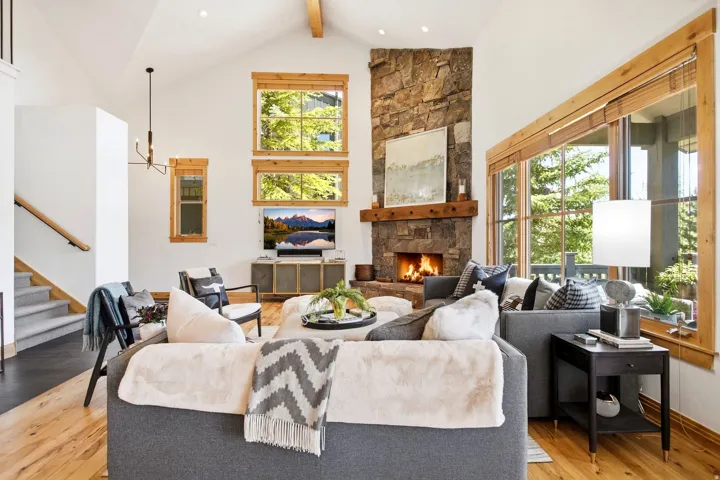 Vaulted ceiling with exposed wood beams, stone-clad fireplace with wood mantel, large windows with natural wood trim, wood-finish flooring, and a carpeted staircase with a wood handrail
