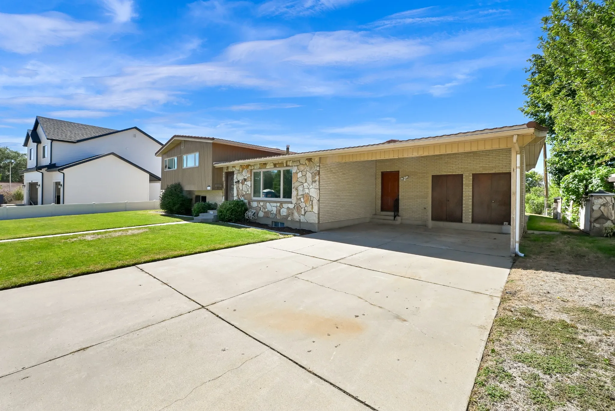 Stone veneer facade, attached carport, expansive concrete driveway, recessed entry, and mature landscaping