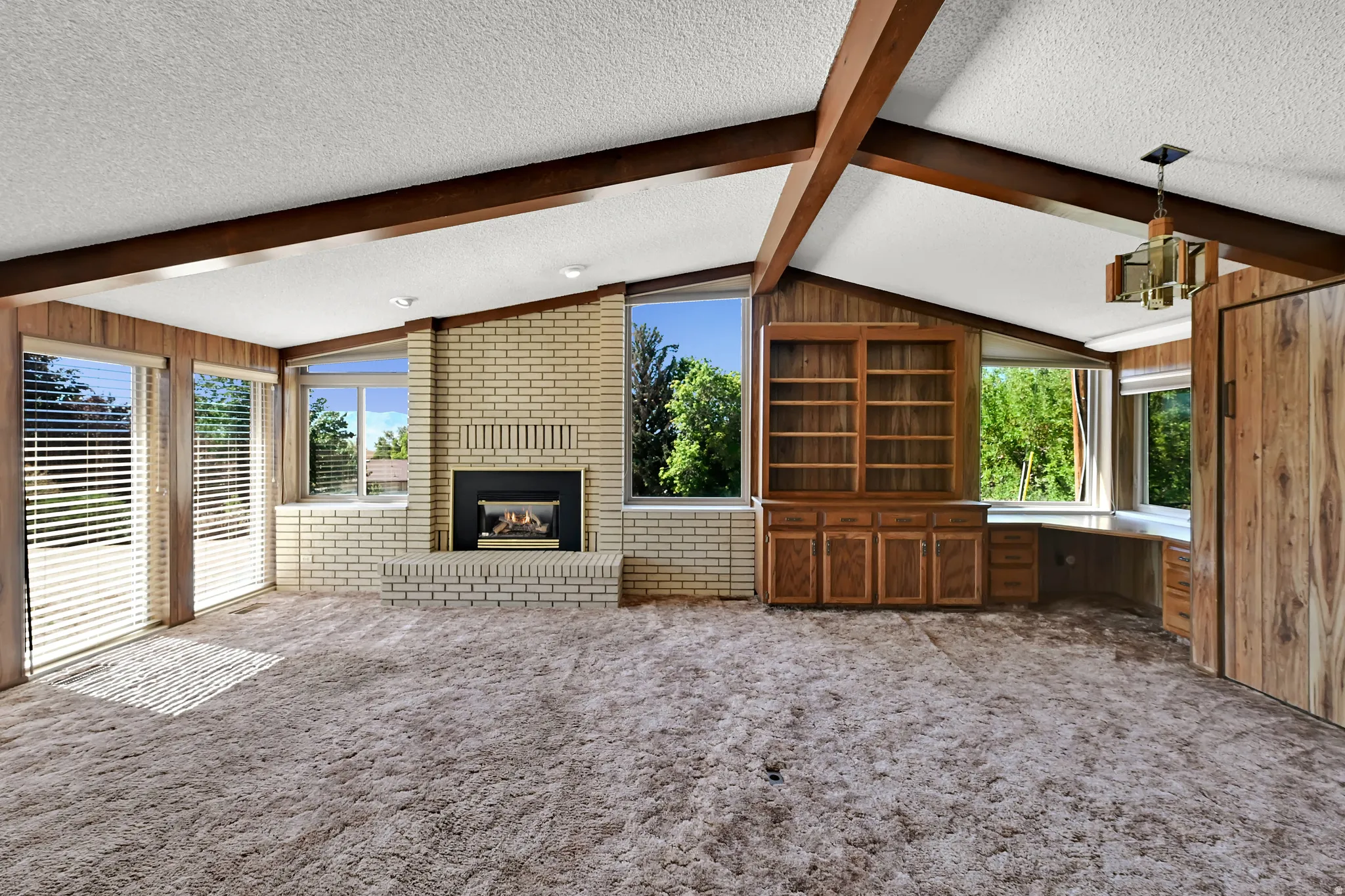 Vaulted ceiling with exposed wood beams, brick fireplace with mantel, built-in desk with cabinetry, built-in shelving, and multiple large windows