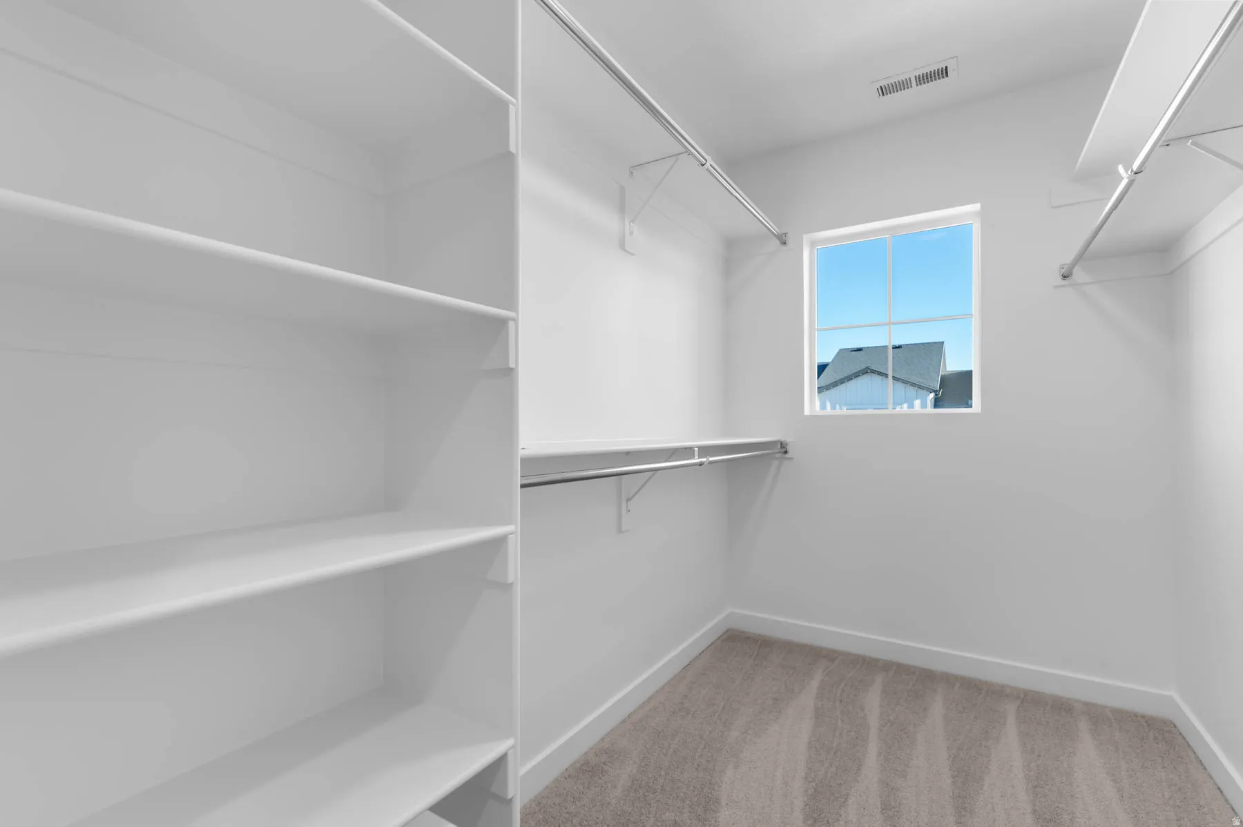 White shelving units, suspended hanging rods, white walls, light-toned carpet flooring, and a window for natural light