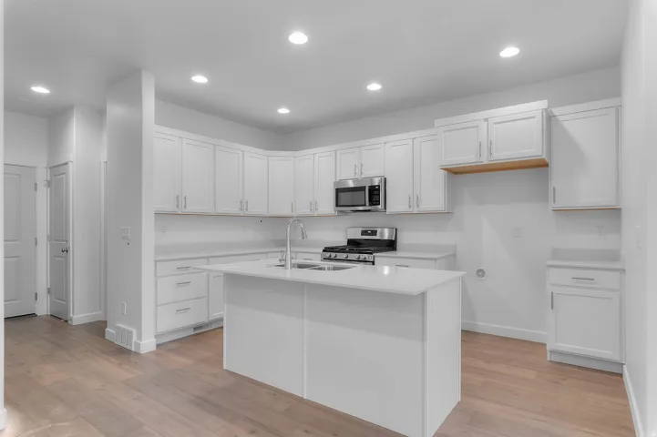 White shaker cabinetry, stainless steel appliances, central island with sink, recessed lighting, and wood-finish flooring