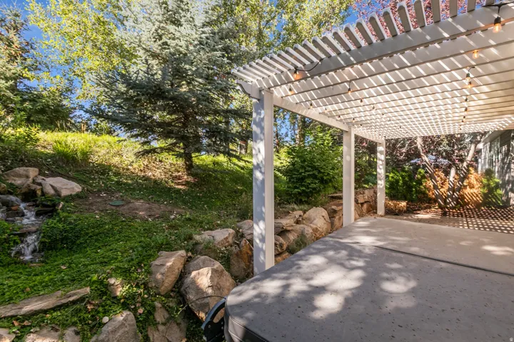 Expansive backyard featuring a white pergola, concrete patio, natural rock landscaping, mature trees, and a water feature