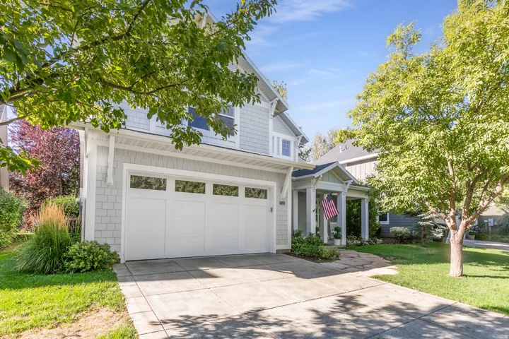 Light gray shingle siding, white trim, attached two-car garage with transom windows, covered front entry porch, and mature trees