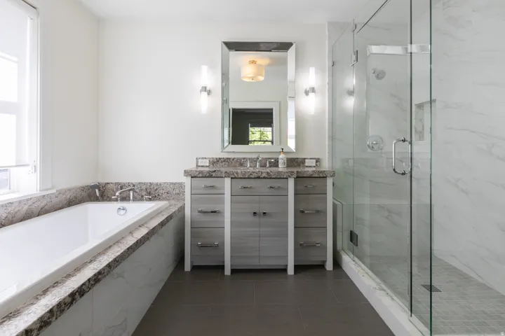 Freestanding soaking tub with a stone surround, glass-enclosed shower with a tiled floor, double vanity with stone countertop, and contemporary wall sconces