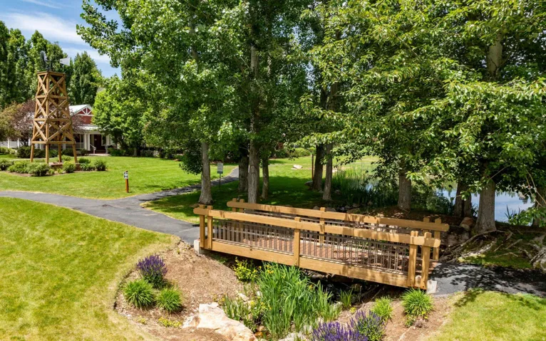 Landscaped grounds featuring a wooden footbridge, paved pathways, mature trees, a water feature, and a decorative windmill structure