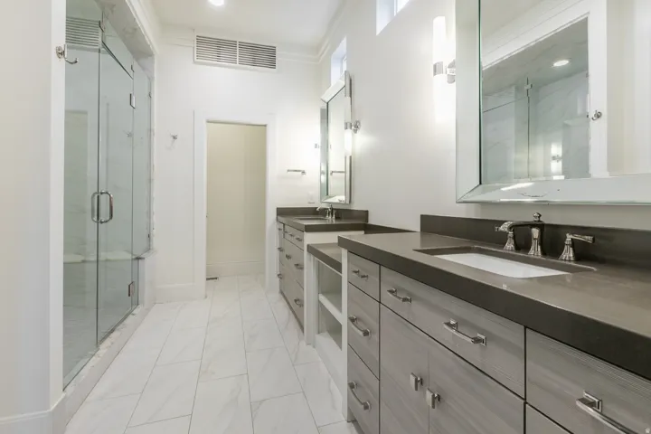 Dual vanity with integrated sinks and dark countertops, glass-enclosed shower, wood-finish cabinetry with polished chrome hardware, and rectangular tile flooring