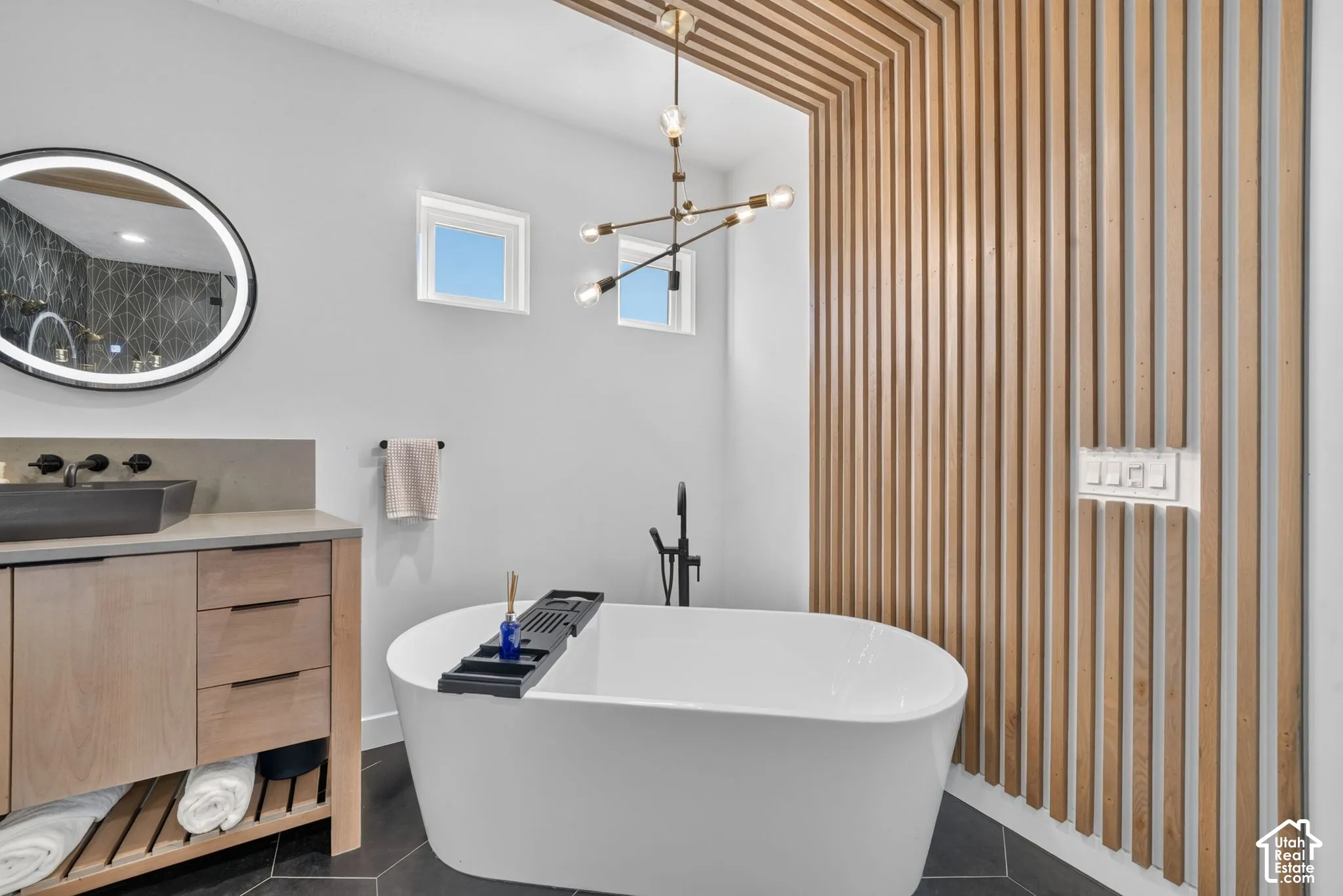 Freestanding bathtub, wood-slat wall feature, dual windows, modern chandelier, and circular illuminated mirror