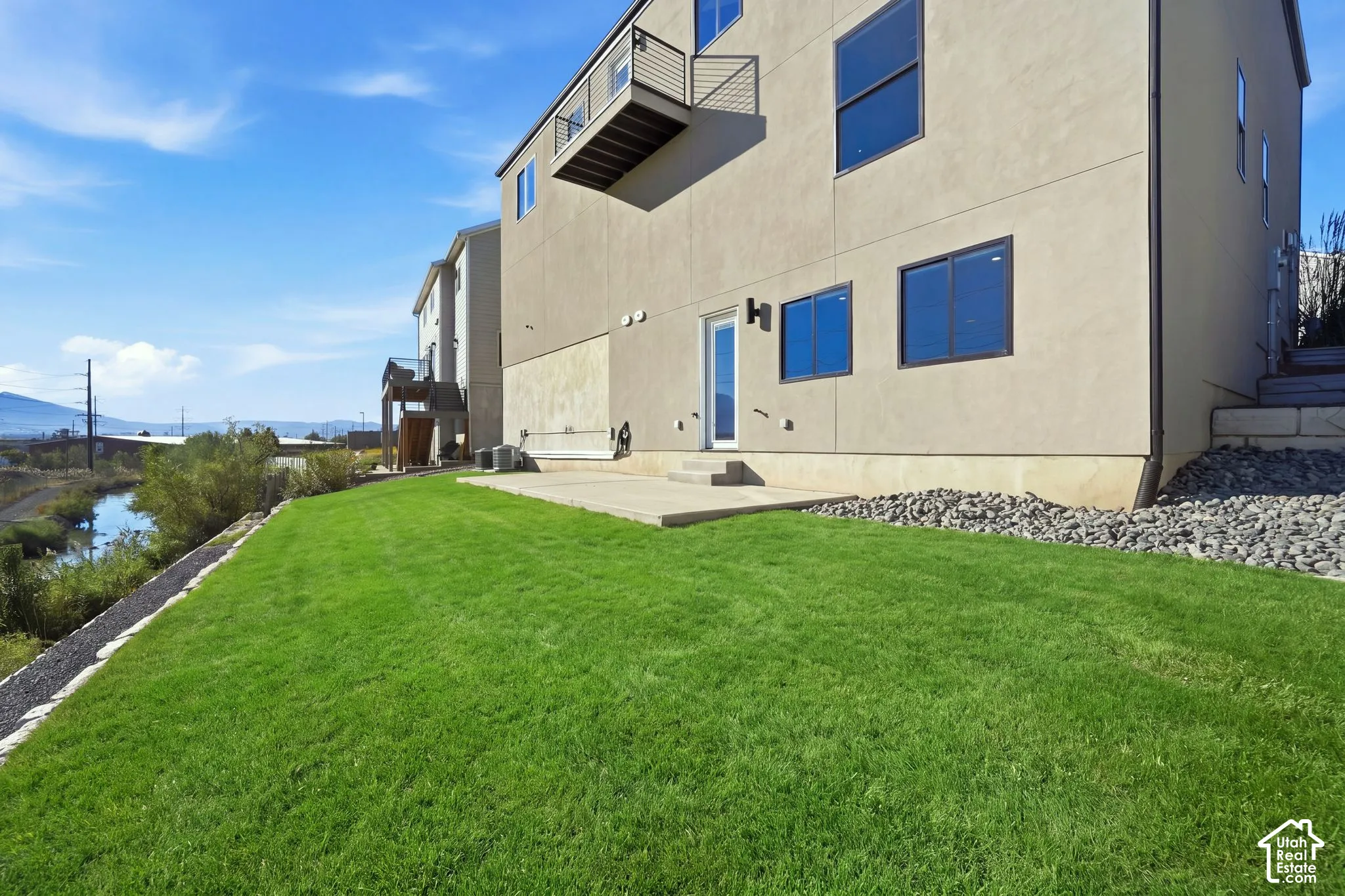 Expansive green lawn with a concrete patio, light-toned exterior siding, black-framed windows, and an upper-level balcony with metal railing