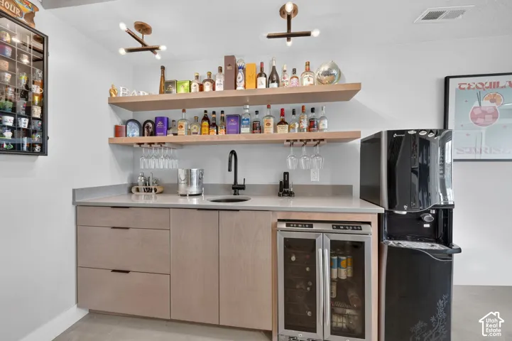 Built-in wet bar featuring a beverage cooler, undermount sink with a matte black faucet, light wood-finish cabinetry, and floating shelves