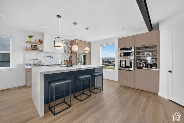 Contemporary kitchen featuring a large center island with black upholstered seating, white cabinetry with waterfall countertops, stainless steel built-in appliances, pendant lighting, and wood-finish flooring throughout the open-concept space
