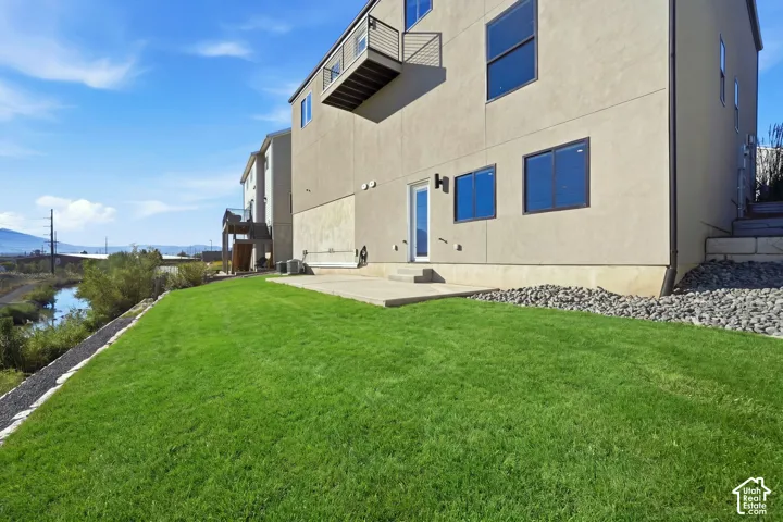 Expansive green lawn with a concrete patio, light-toned exterior siding, black-framed windows, and an upper-level balcony with metal railing