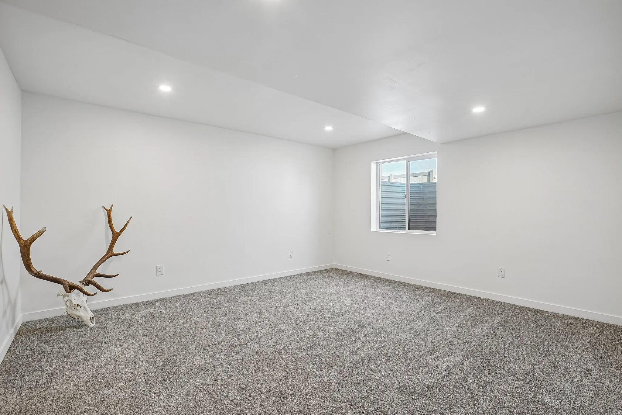 Recessed lighting, light-colored walls, gray carpeting, and a single window with horizontal blinds