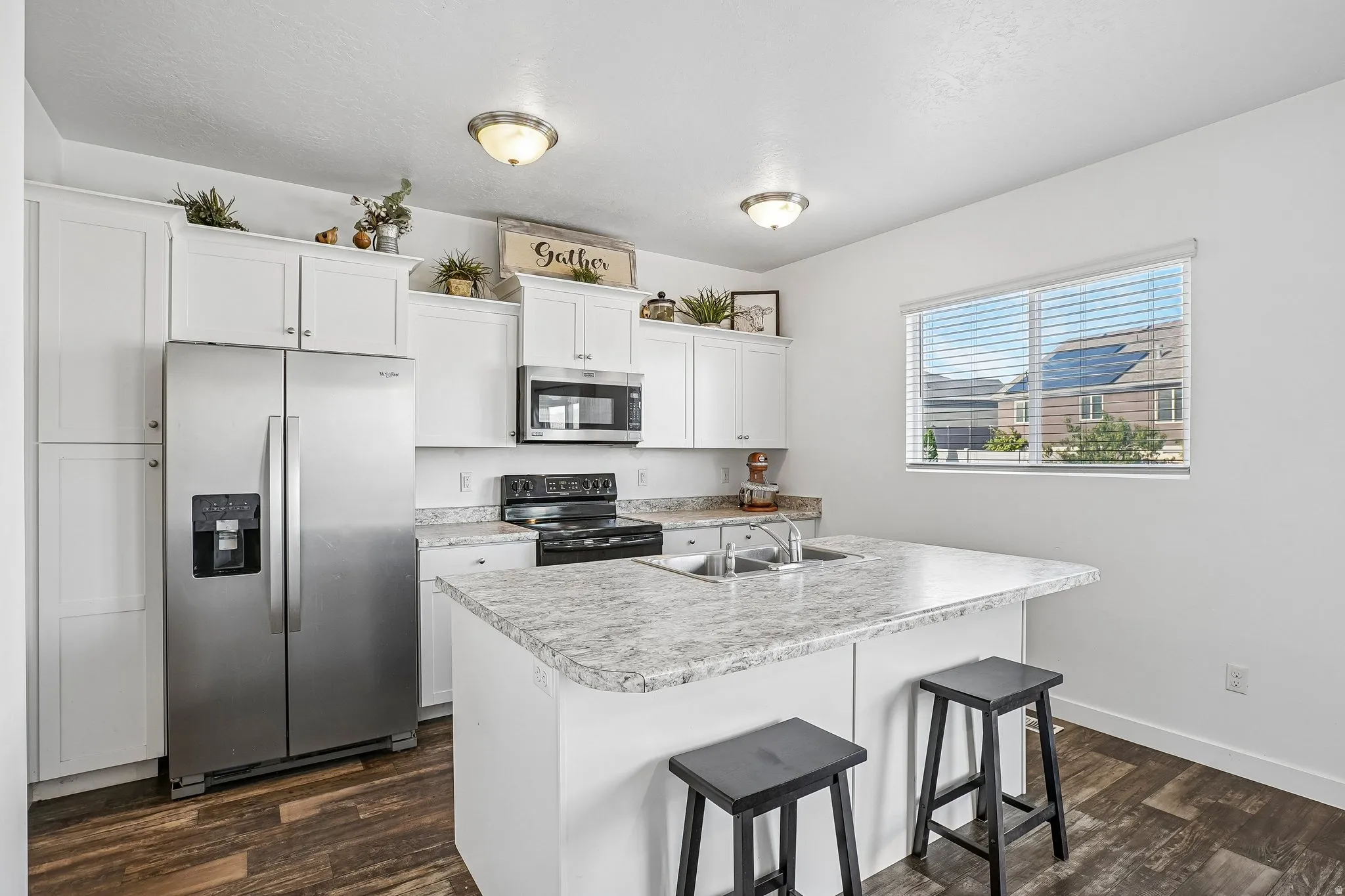 Kitchen featuring white cabinetry, stainless steel appliances, an island with an integrated sink, wood-finish flooring, and a window with blinds