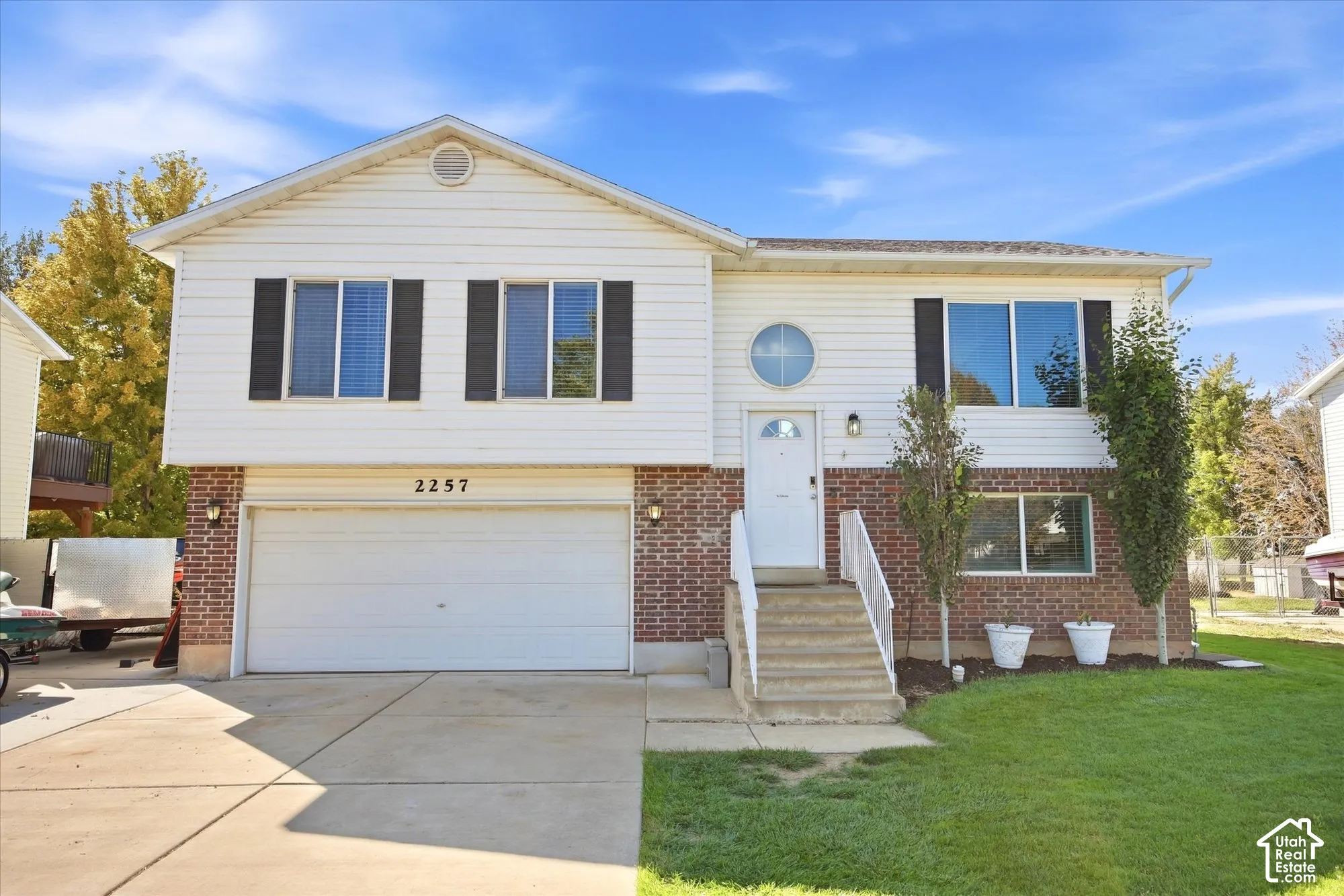 Split-level exterior featuring white siding and brick accents, an integrated two-car garage, multiple windows with dark shutters, a front entry with a circular window, and a concrete driveway