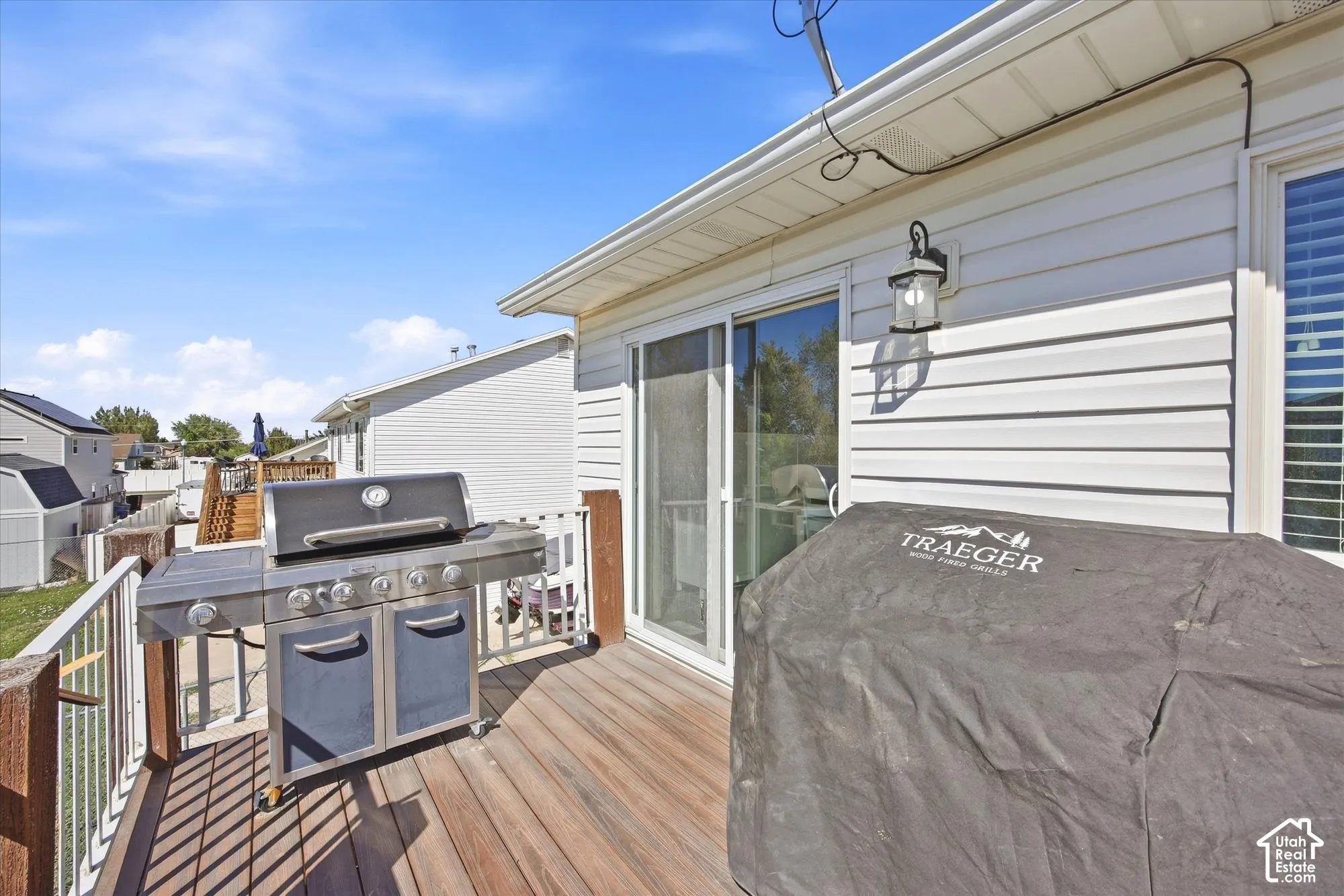 Expansive wood-finish deck with white railing, featuring a sliding glass door and exterior wall lantern on vinyl siding