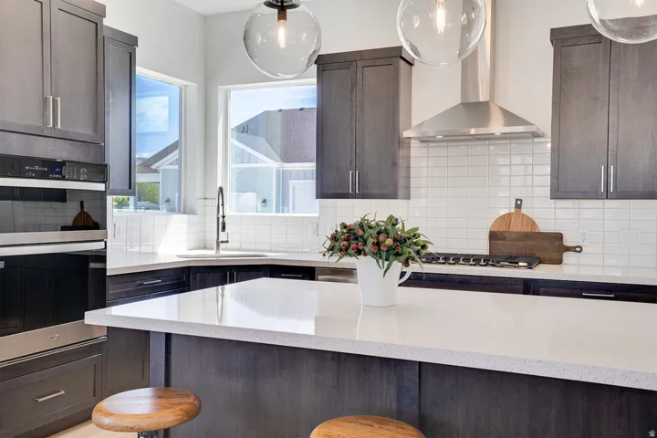 Modern kitchen featuring dark wood-finish cabinetry, light-toned countertops, a stainless steel range hood, built-in double ovens, and a white subway tile backsplash
