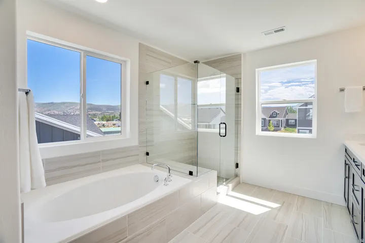 Spacious bathroom featuring a soaking tub, glass-enclosed shower, light-toned tile flooring, and multiple windows