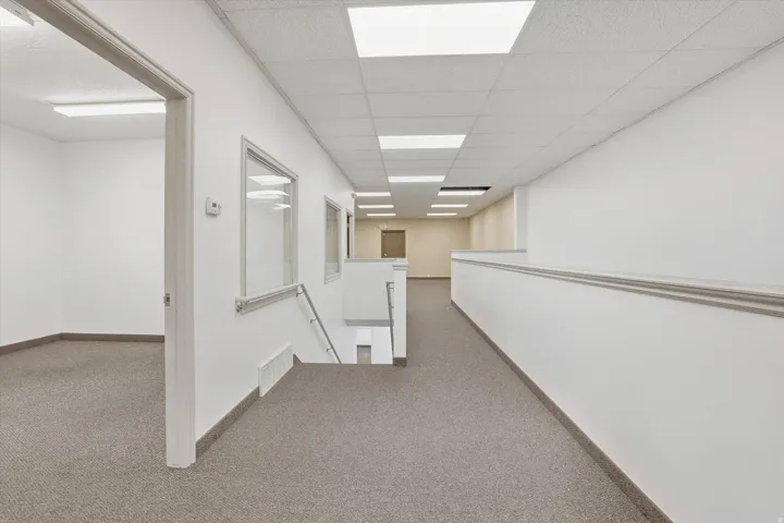 Carpeted hallway with white walls, recessed fluorescent lighting, and a partial glass partition