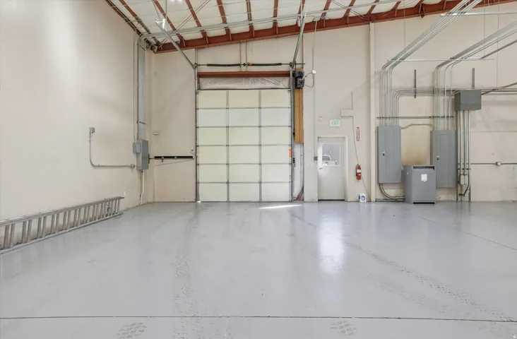 High-clearance roll-up door, polished concrete flooring, exposed ceiling trusses, interior entry door, and multiple electrical panels