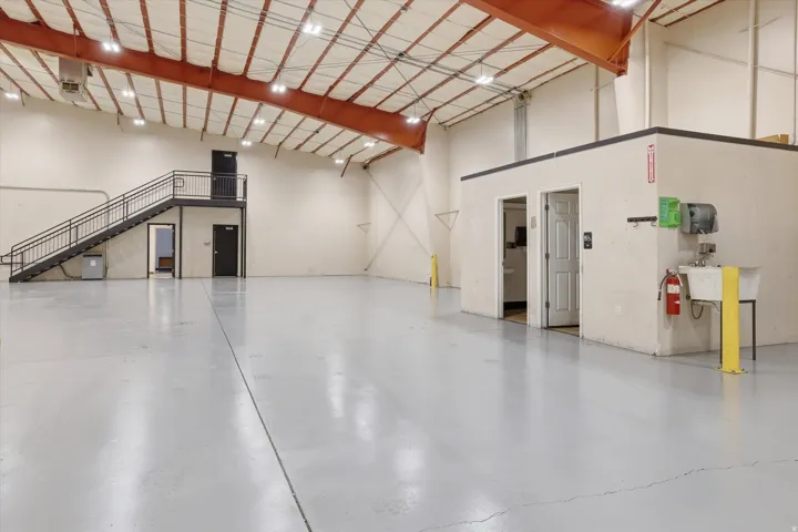 Expansive warehouse space with high ceilings, polished concrete flooring, a built-in office structure, and an interior staircase providing access to an upper-level platform