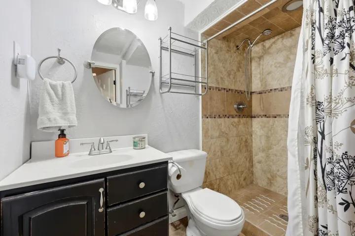 Tiled shower with decorative accent band, dark wood-finish vanity, white countertop with integrated sink, and wall-mounted mirror