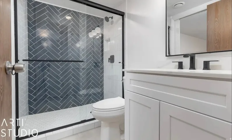 Glass-enclosed shower with herringbone tile, white vanity with integrated sink, black-framed mirror, and matte black fixtures