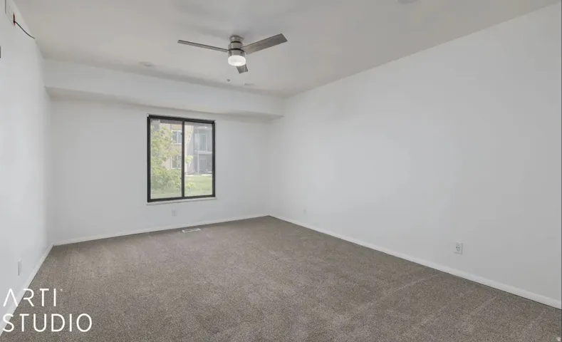 Spacious room featuring neutral carpeting, white walls, a large window with black trim, and a ceiling fan with integrated lighting