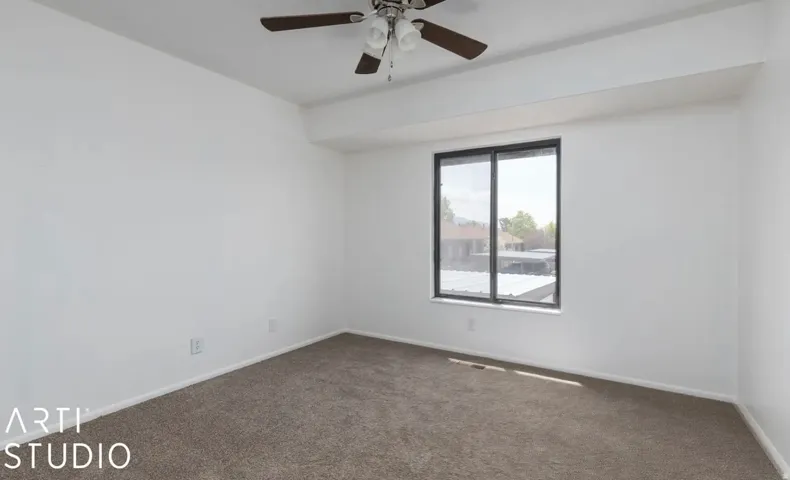 Neutral wall paint, ceiling fan with light fixture, window with dark frame, neutral tone carpet flooring, white trim