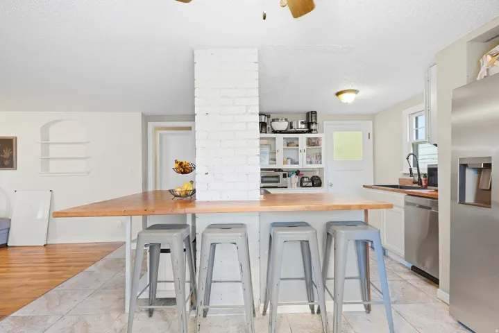 Kitchen featuring a wood-finish countertop island, white brick accent wall, white cabinetry with glass inserts, stainless steel appliances, and tile flooring
