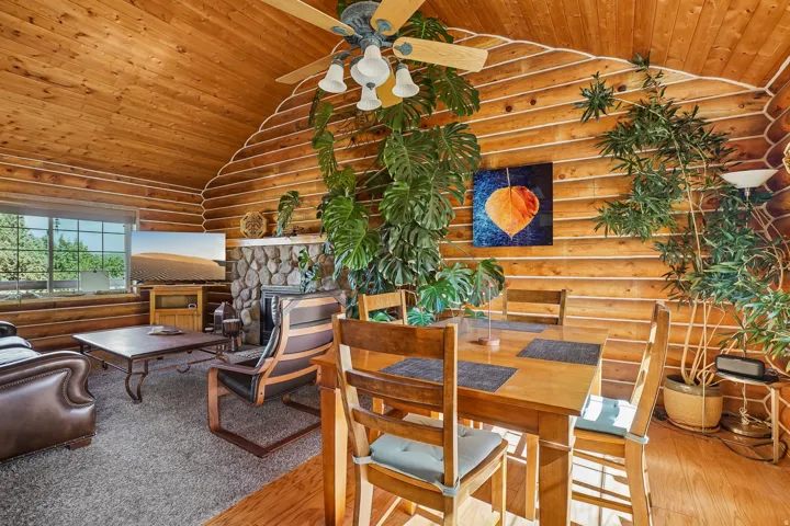 Vaulted wood-paneled ceiling with a central fan, log-style wood walls, stone fireplace surround, wood-finish flooring, and a large window