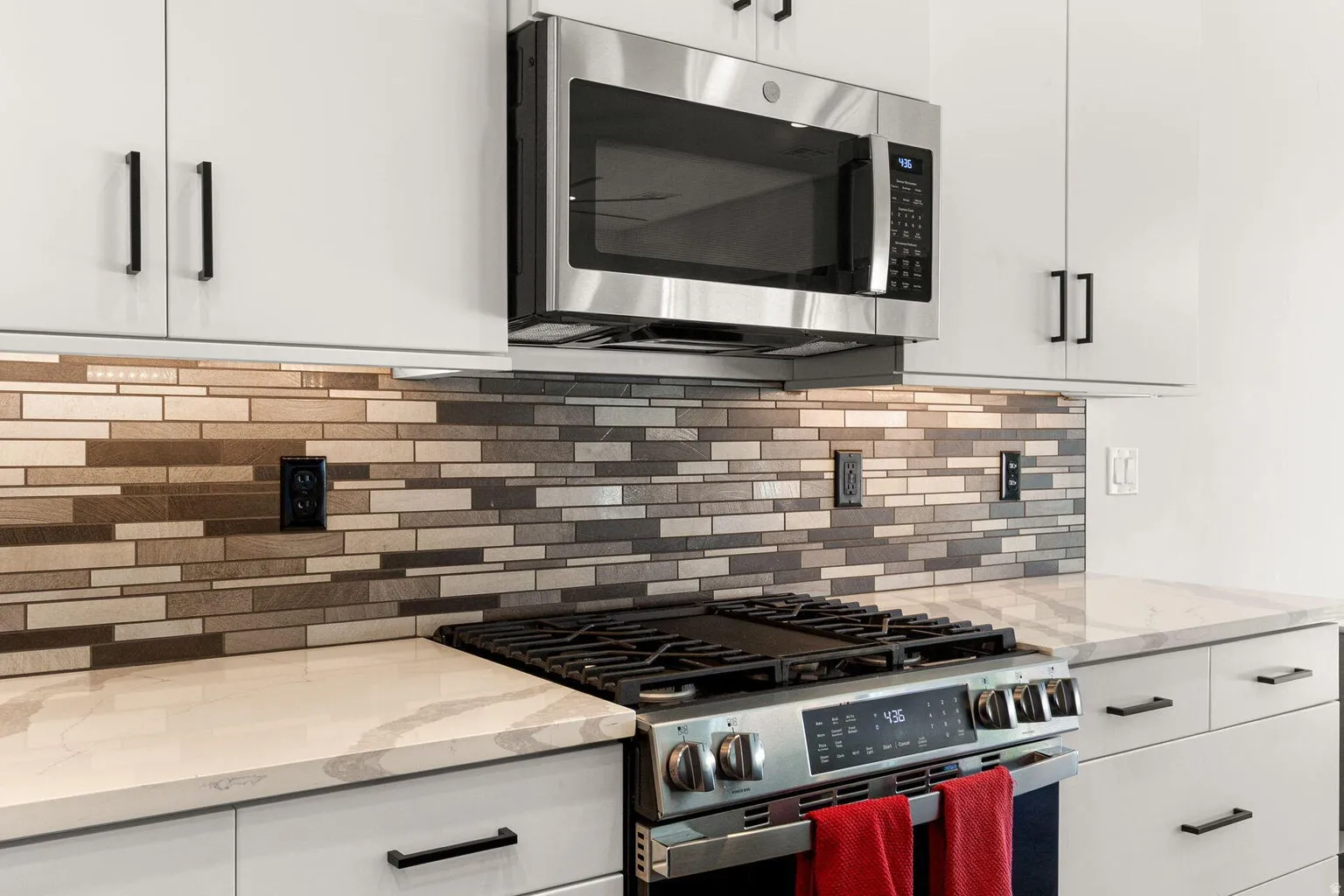 Stainless steel microwave and gas range, white cabinetry with matte black hardware, mosaic tile backsplash, and light-toned countertops