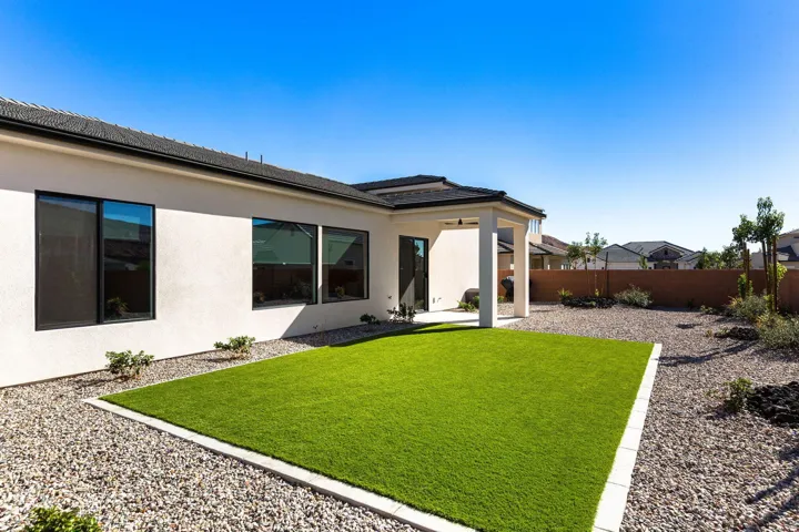 Expansive backyard featuring a rectangular artificial turf lawn, white stucco exterior, black-framed windows, and a covered patio area