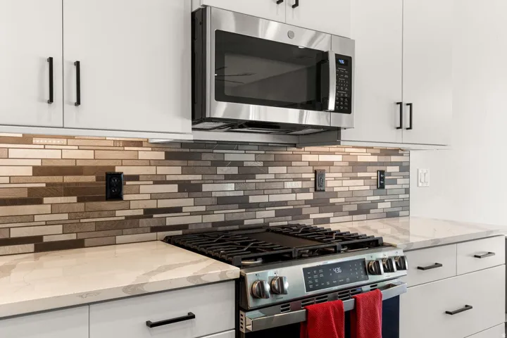 Stainless steel microwave and gas range, white cabinetry with matte black hardware, mosaic tile backsplash, and light-toned countertops