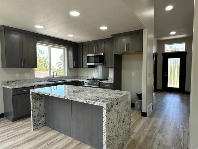 Kitchen featuring dark shaker-style cabinetry, stainless steel appliances, a center island with a waterfall countertop, and wood-finish flooring