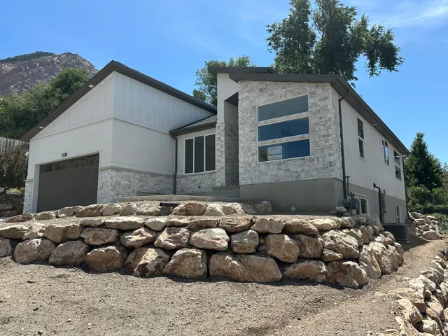 Modern exterior featuring stone veneer accents, stucco siding, a two-car garage, multiple large windows, and a natural rock retaining wall