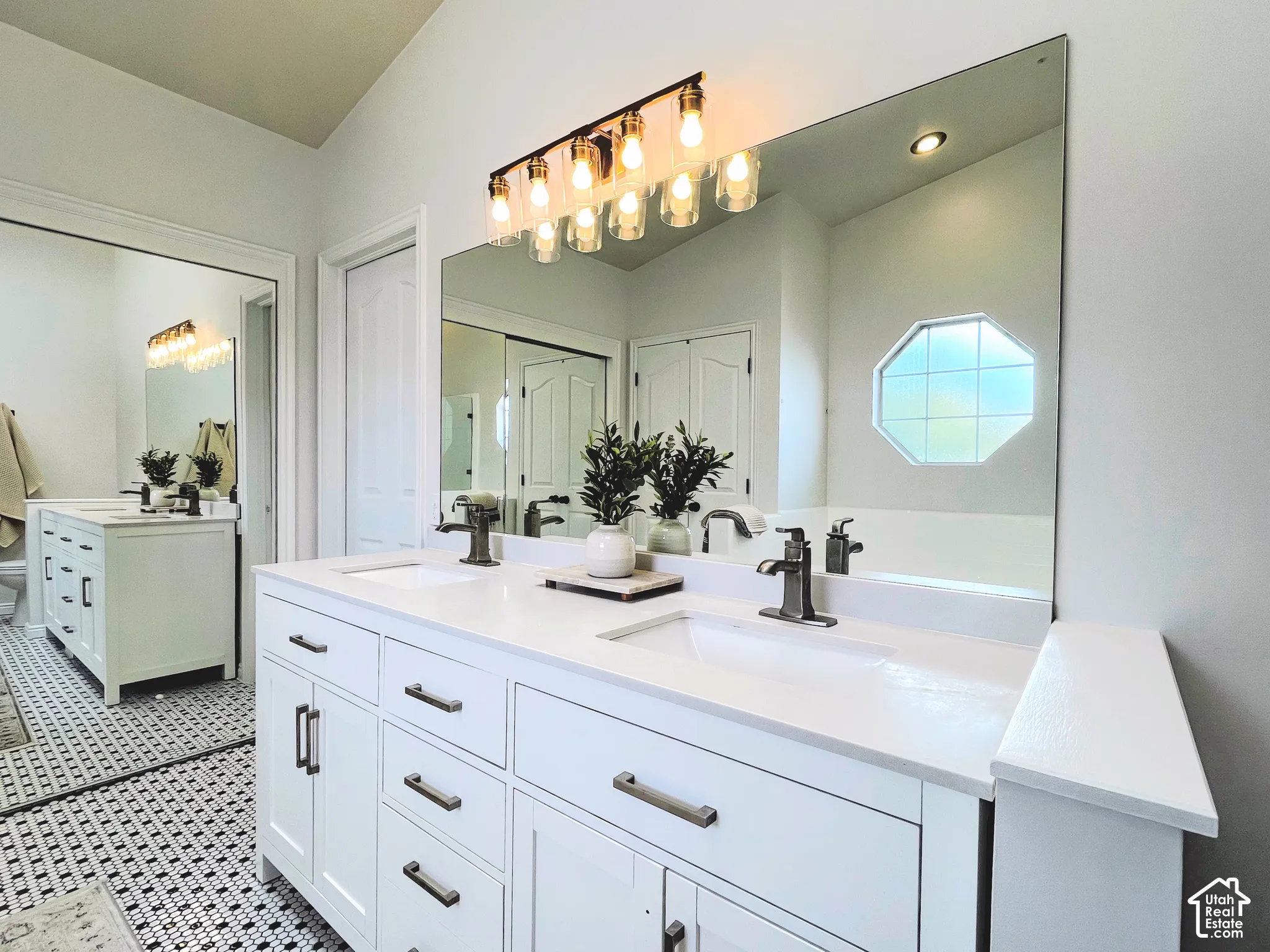 Dual white vanities with integrated sinks, brushed nickel fixtures, a large frameless mirror, and a black and white mosaic tile floor