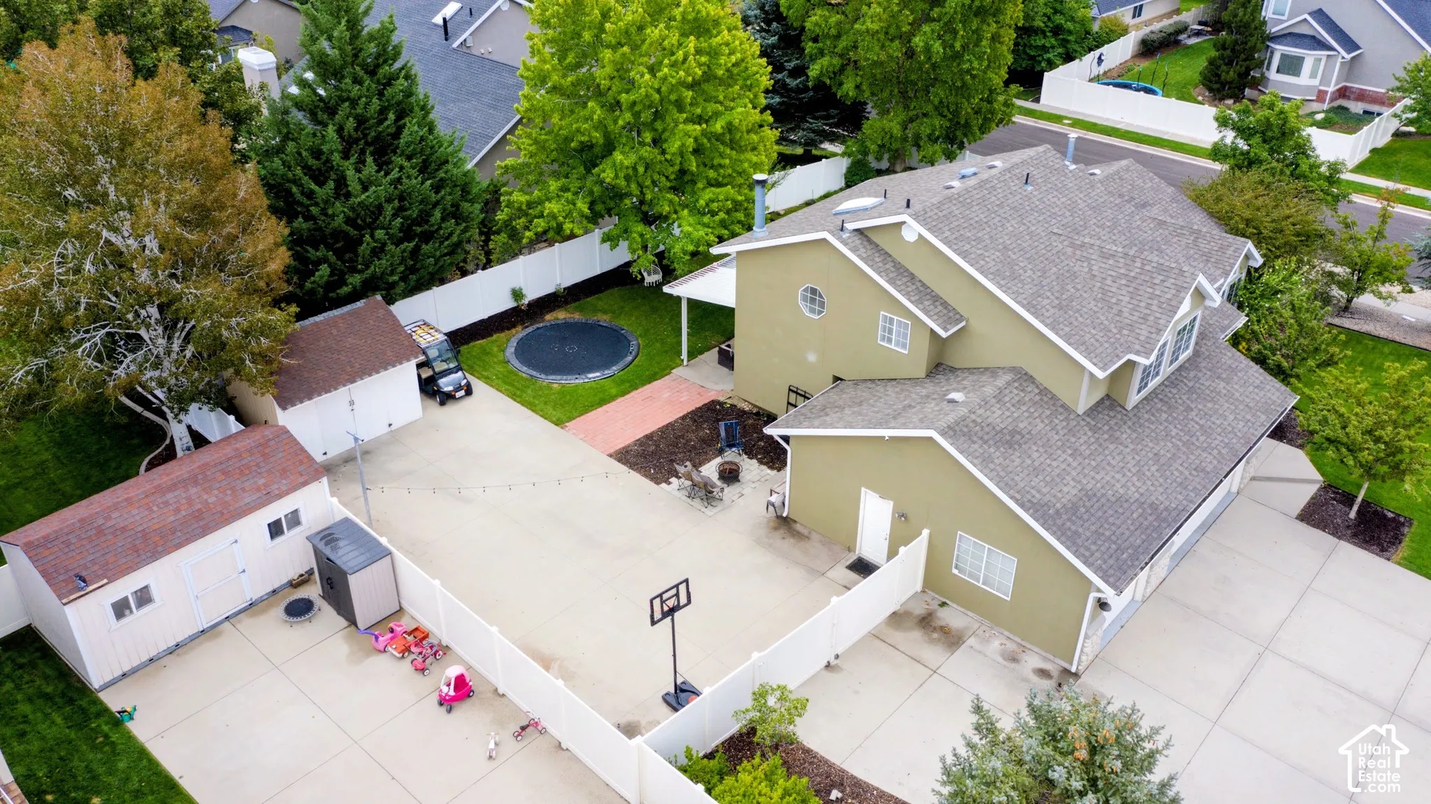 Expansive concrete driveway, two detached storage sheds, fenced backyard with a lawn area, and mature trees providing shade