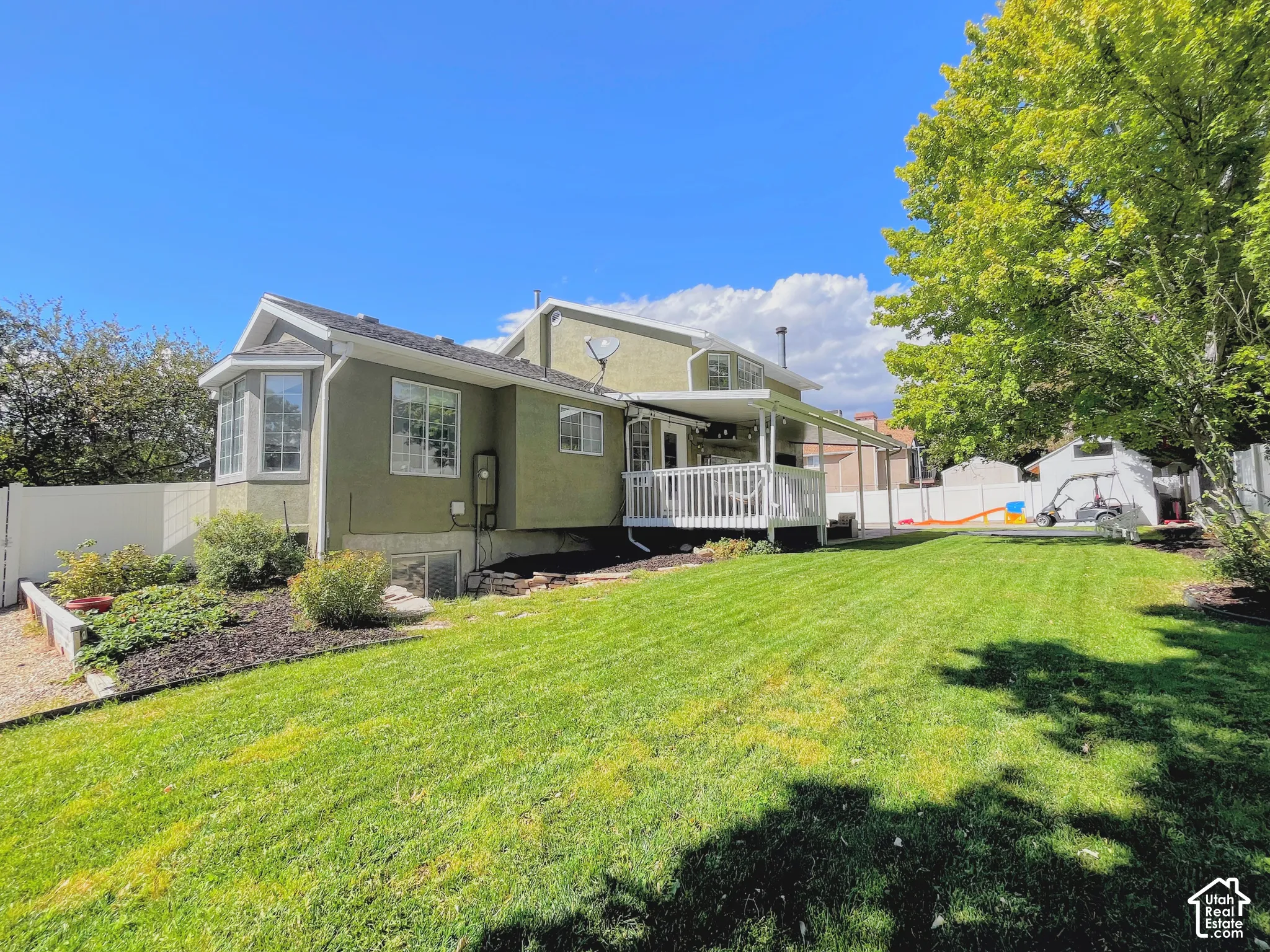 Expansive green lawn, a covered white deck, exterior bay window, stucco exterior, and mature landscaping